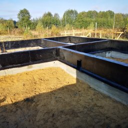 Forma Budownictwo Paweł Dąbrowski - Zabezpieczone fundamenty domu jednorodzinnego z widoczną hydroizolacją w postaci czarnej powłoki bitumicznej, piasek wewnątrz wykopu, drewniane elementy szalunku w tle.