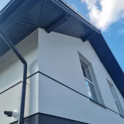 Fragment elewacji domu z widocznymi oknami, orynnowaniem i podbitką dachową w kolorze grafitowym, na tle błękitnego nieba z białymi chmurami. Dolna część elewacji w kolorze ciemnoszarym, górna...