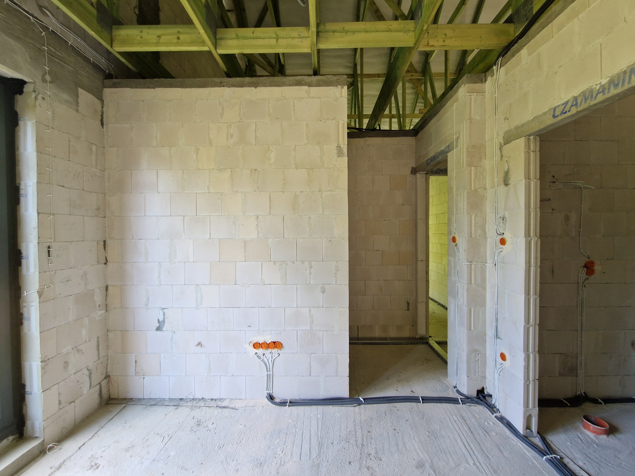 Wnętrze surowego domu w trakcie budowy, ściany z białych bloczków, widoczne instalacje elektryczne w peszlach, drewniana konstrukcja dachu.