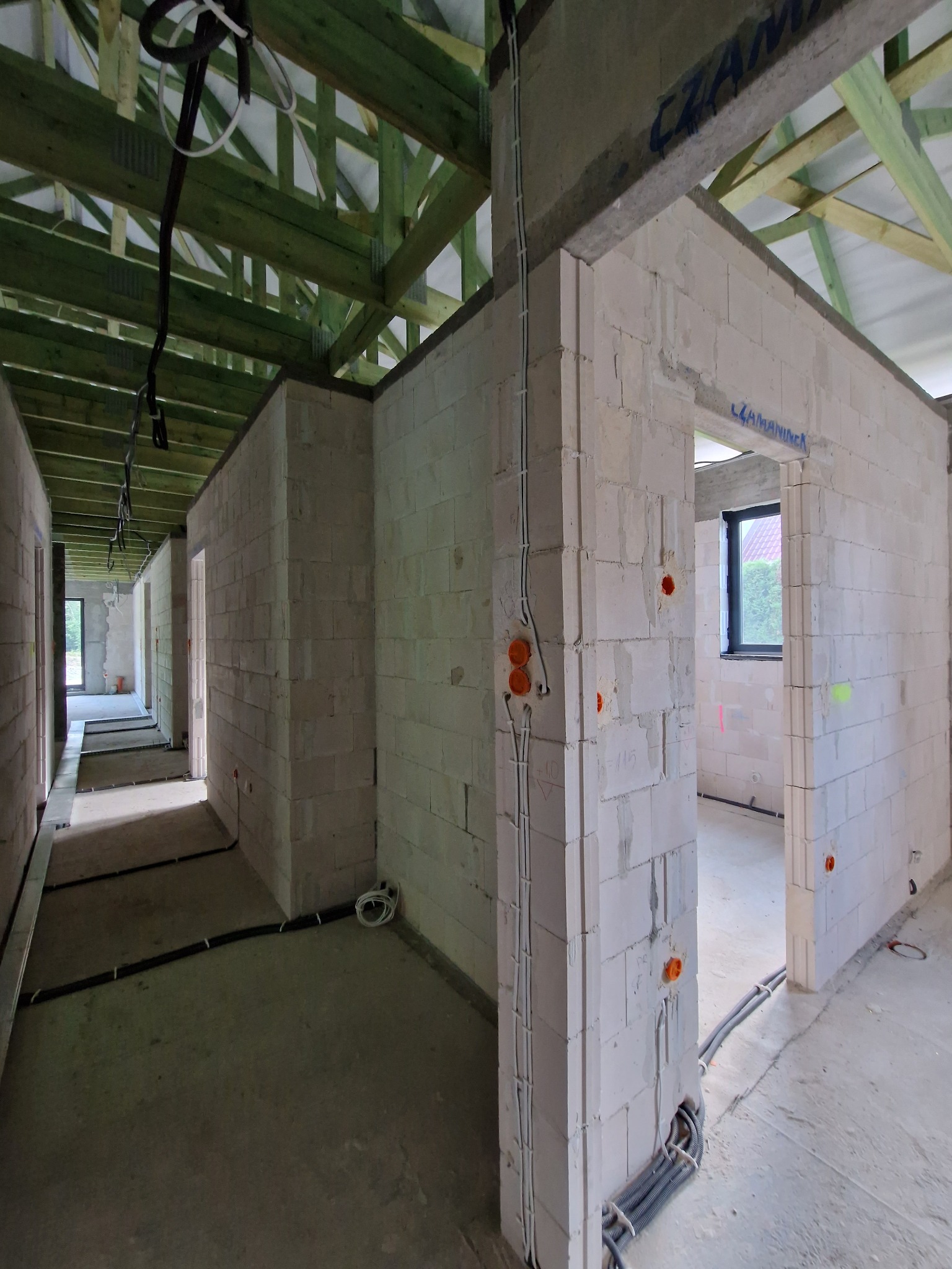 Wnętrze domu w trakcie budowy, widoczne ściany z białych bloczków, drewniana konstrukcja dachu, instalacja elektryczna w ścianach i na podłodze, surowy betonowy podkład.