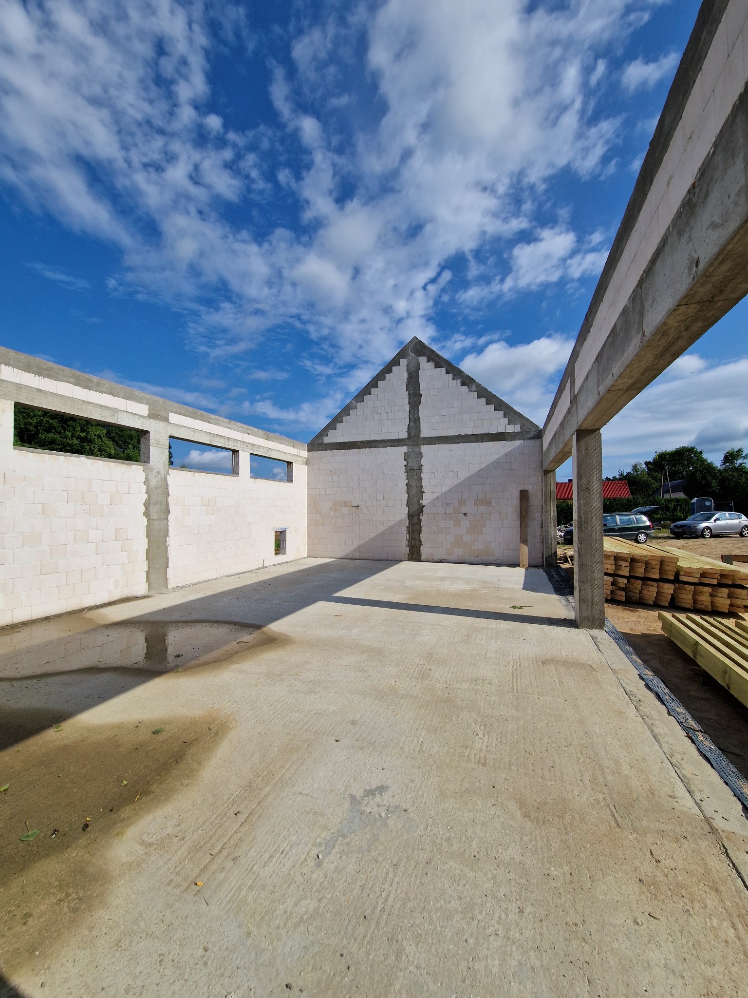 Stan surowy otwarty budynku z białych bloczków, widoczne ślady po deszczu na betonowej posadzce, w tle błękitne niebo z chmurami, przygotowane drewniane bale.