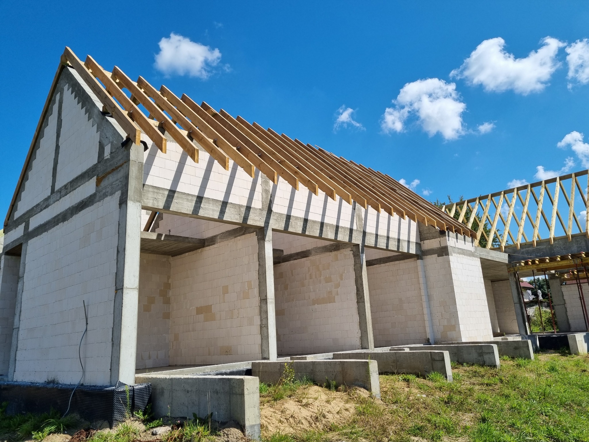 Surowy stan domu murowanego z betonu komórkowego, widoczne ściany, słupy konstrukcyjne i więźba dachowa, na tle błękitnego nieba z chmurami.