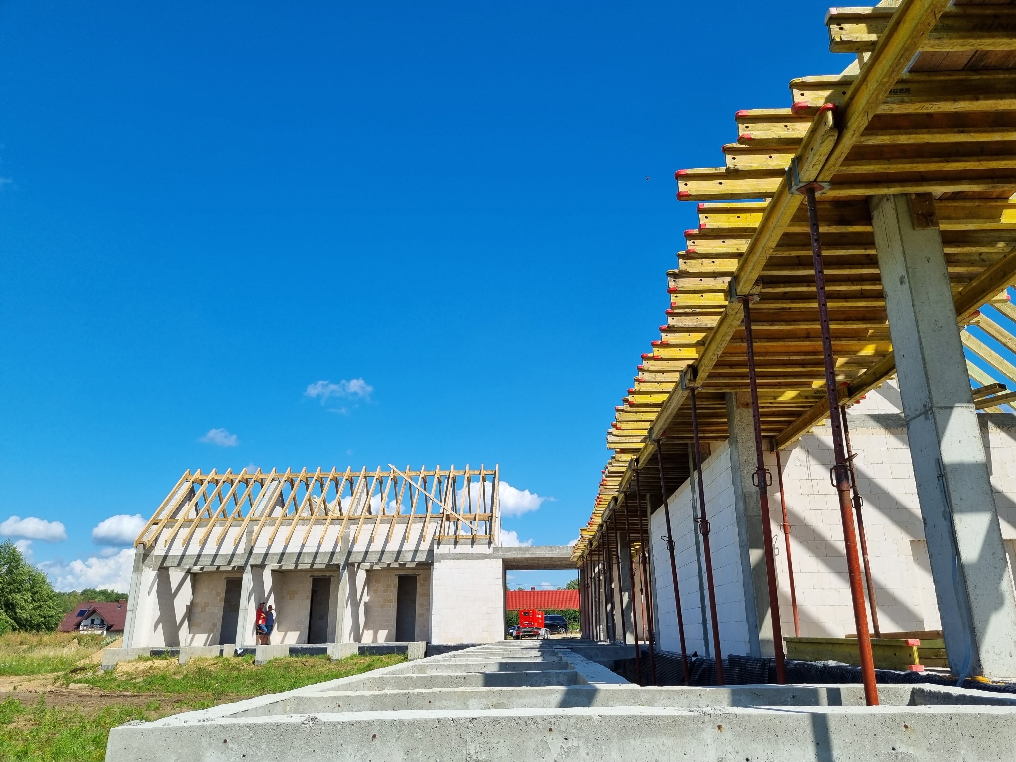Wznoszenie budynku z białych bloczków, widoczne szalunki stropowe wsparte na stalowych podporach, w tle błękitne niebo i częściowo ukończony dach z drewnianą konstrukcją.