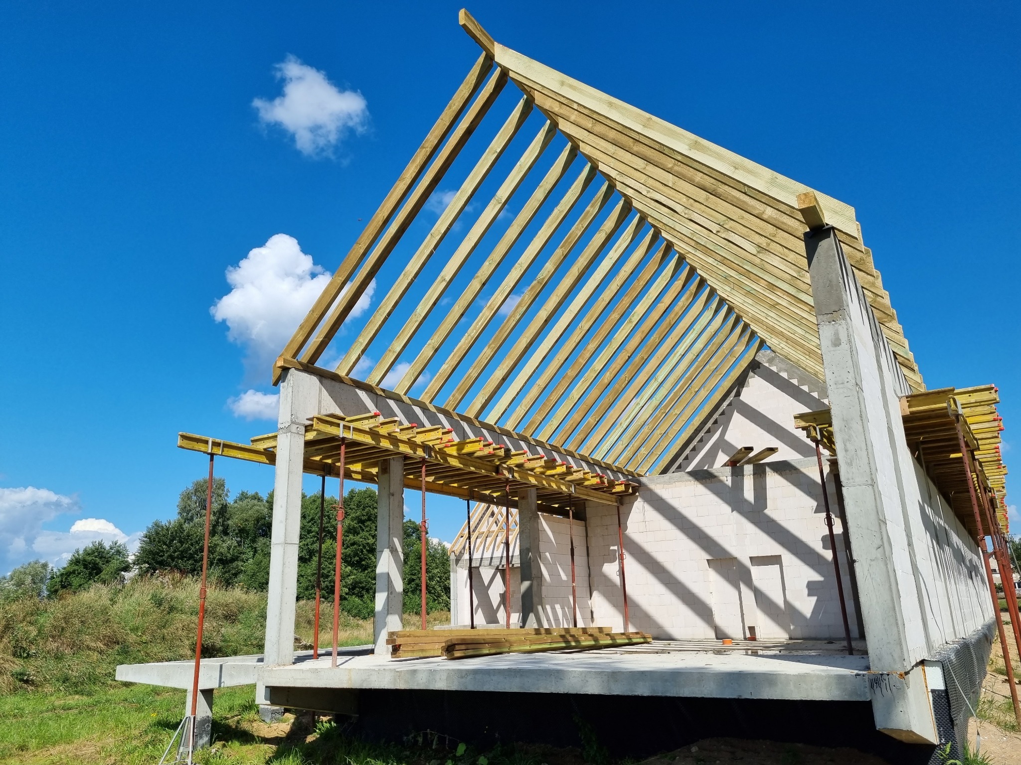 Surowy stan domu jednorodzinnego z betonu komórkowego z widoczną konstrukcją drewnianą dachu na tle błękitnego nieba z chmurami.