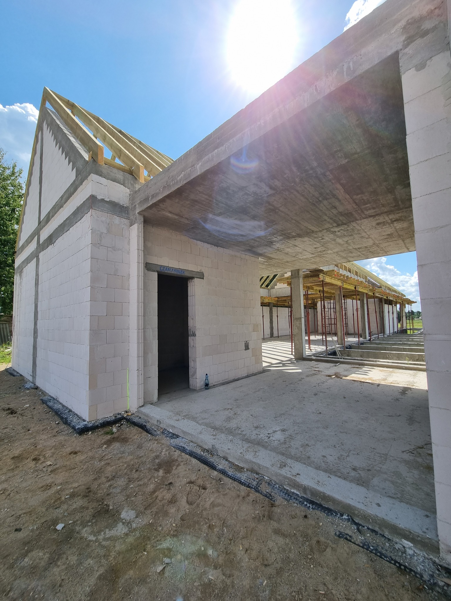 Surowy stan budynku z betonu komórkowego i drewnianą konstrukcją dachu w trakcie budowy, widoczne szalunki stropu, słoneczny dzień.