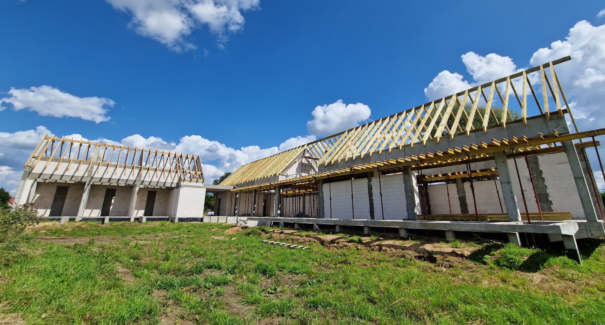 Budowa domu w stanie surowym otwartym: ściany z betonu komórkowego, drewniana konstrukcja dachu w trakcie montażu, widoczne podpory szalunkowe, błękitne niebo z chmurami w tle.