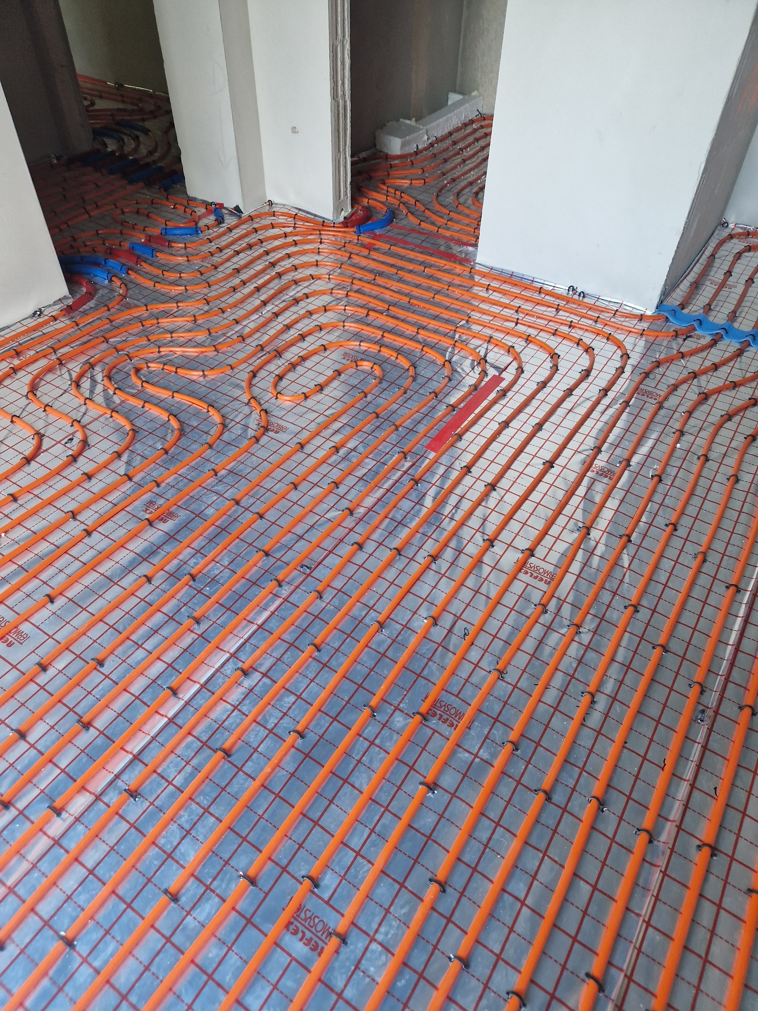 Pomarańczowe rury ogrzewania podłogowego ułożone w pętle na srebrnej folii refleksyjnej z siatką, mocowane czarnymi klipsami w surowym wnętrzu budynku w trakcie budowy.