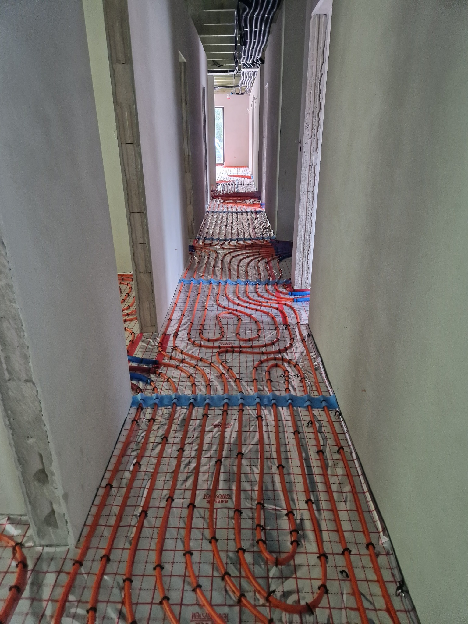 Pomieszczenie w stanie surowym z widoczną instalacją ogrzewania podłogowego z pomarańczowych rurek na folii z siatką, zamocowanych niebieskimi klipsami, ściany w szarym tynku, widok w perspektywie...