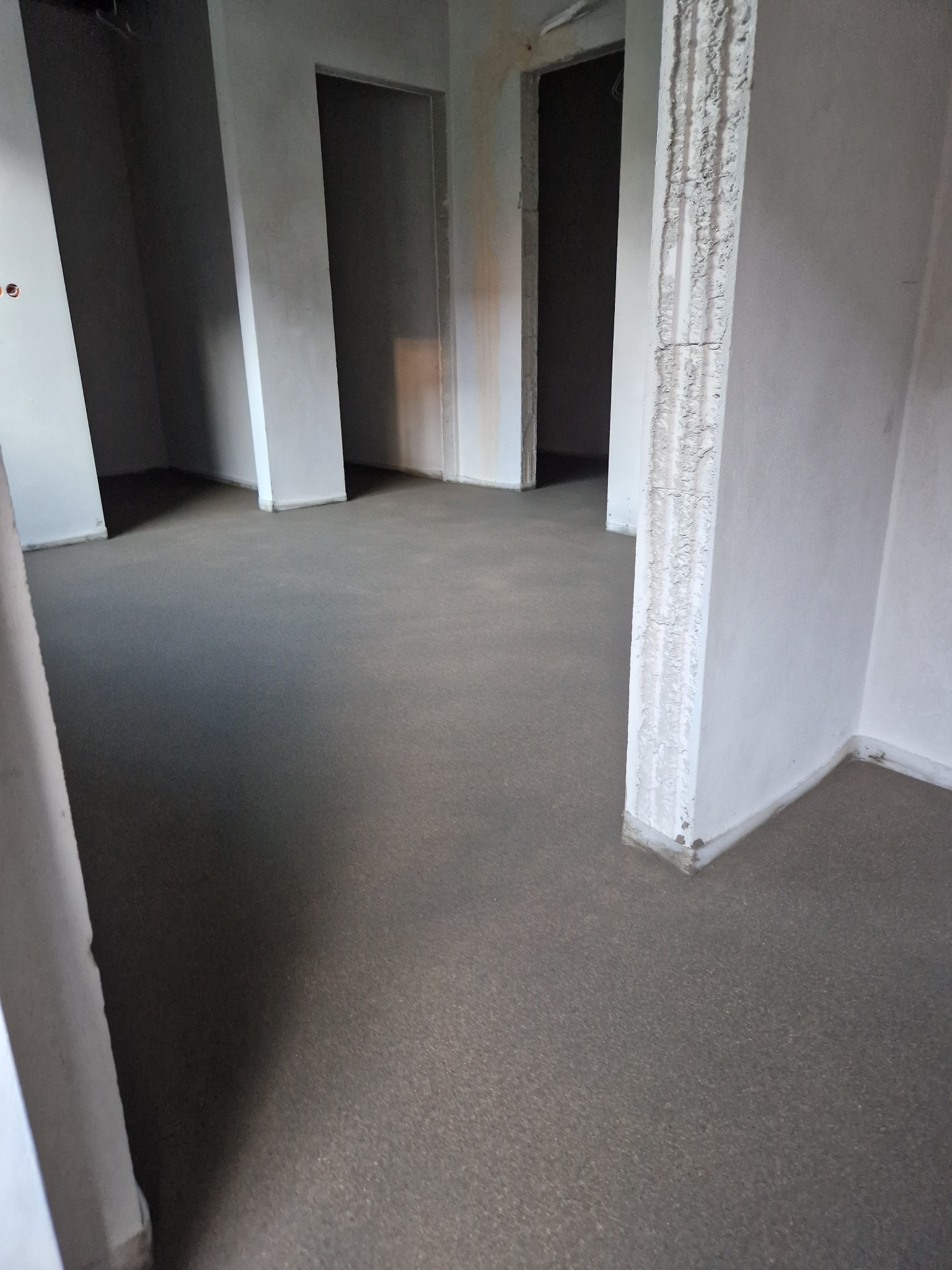 Wnętrze domu w trakcie budowy z wylaną, gładką posadzką, surowe ściany i widoczne surowe otwory drzwiowe.