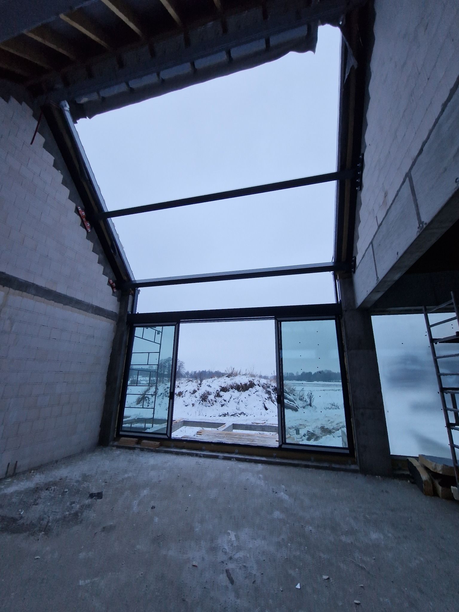 Wnętrze surowego domu w Białymstoku z widocznymi ścianami z betonu komórkowego, dużym oknem przesuwnym z widokiem na zaśnieżony krajobraz i elementami konstrukcji dachu.
