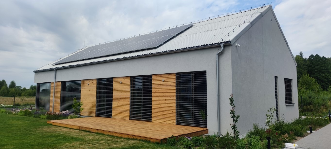 Nowoczesny dom jednorodzinny z panelami słonecznymi na dachu, elewacja w odcieniach szarości, drewniane elementy dekoracyjne na fasadzie, drewniany taras, zadbany trawnik i rabaty kwiatowe.