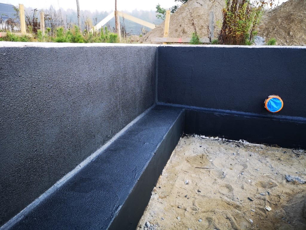 Zabezpieczone hydroizolacją fundamenty domu w trakcie budowy, widoczna warstwa izolacyjna w kolorze czarnym oraz otwór kanalizacyjny zabezpieczony niebieską folią.
