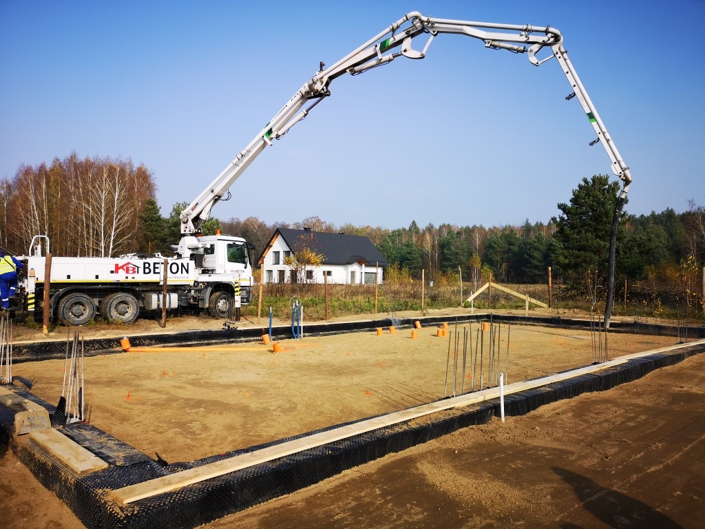 Wylewanie betonu na fundamenty domu jednorodzinnego za pomocą pompy do betonu KB Beton. Widoczne zbrojenia, szalunki i izolacja pionowa fundamentów. W tle dom w stanie surowym otwartym.