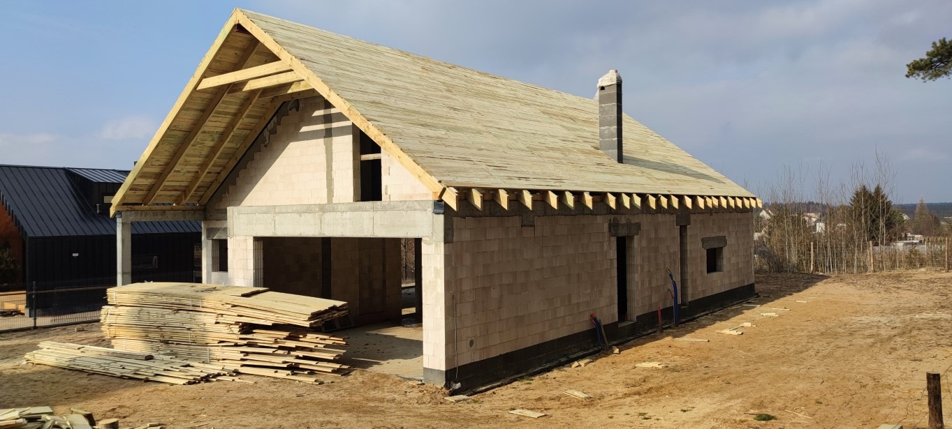 Surowy stan domu murowanego z pustaków, widoczna konstrukcja dachu, komin, izolacja fundamentów i stos desek na placu budowy.