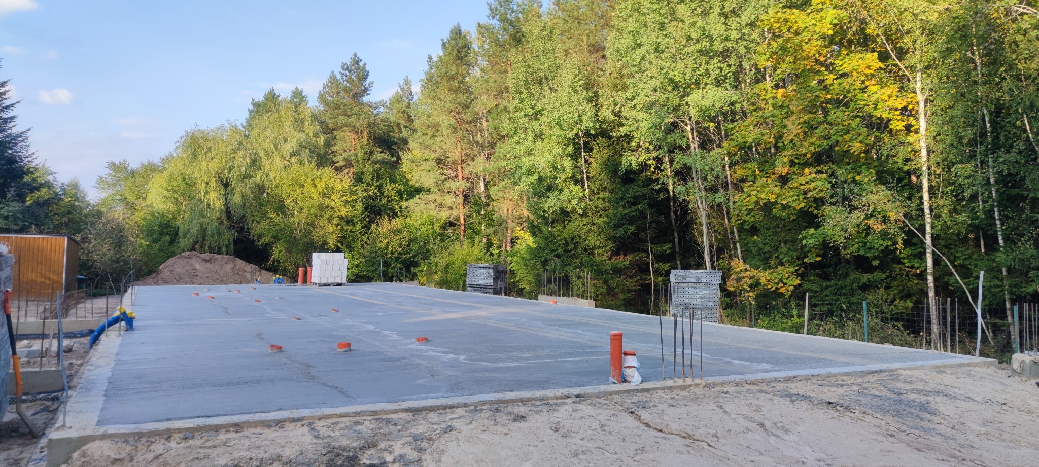 Wylana płyta fundamentowa z wystającymi rurami i elementami konstrukcyjnymi, widok na pobliski las.