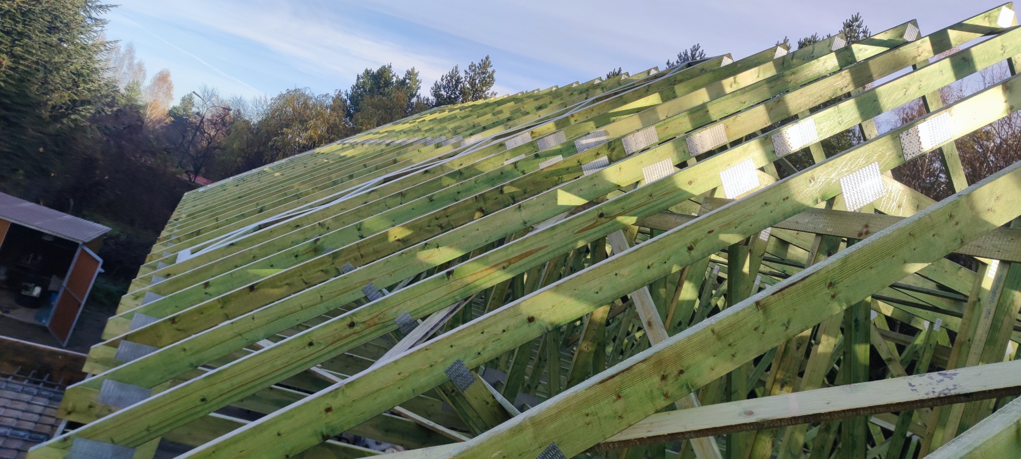 Konstrukcja drewniana dachu w trakcie budowy, widoczne impregnaty i metalowe łączniki, tło stanowią drzewa i błękitne niebo.