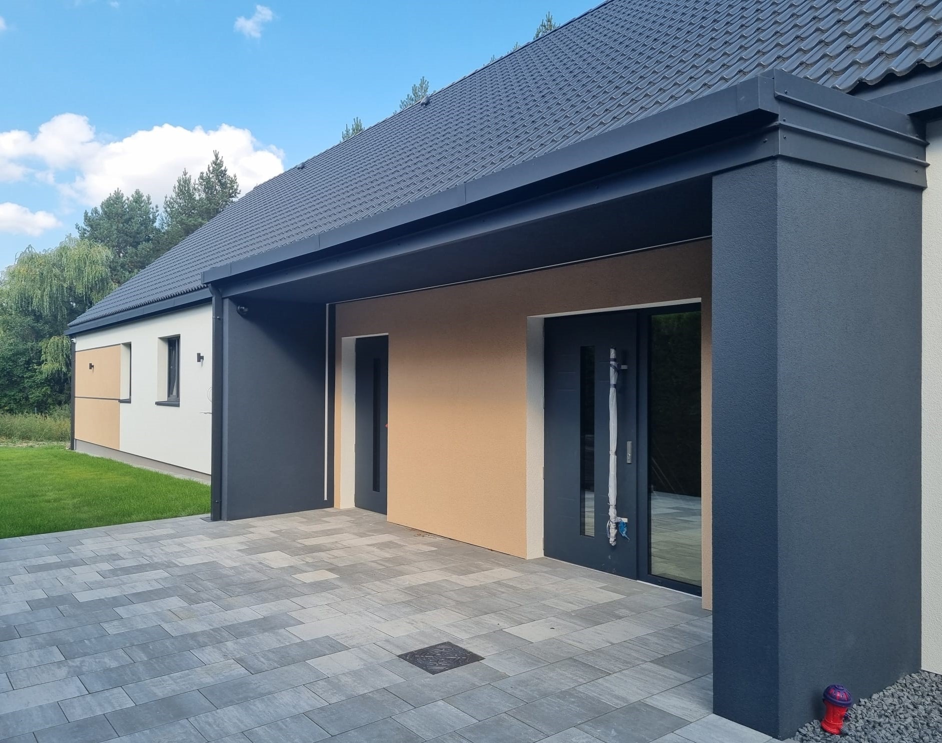 Nowoczesny dom jednorodzinny z ciemnoszarą stolarką okienną i drzwiową, elewacją w odcieniach bieli, beżu i szarości, oraz brukowanym podjazdem z odpływem liniowym. Na pierwszym planie czerwony...