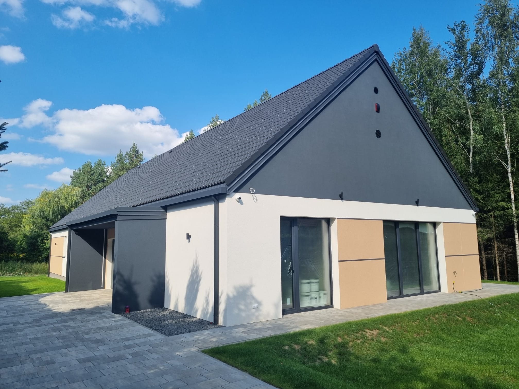 Nowoczesny dom jednorodzinny z ciemnoszarą dachówką i elewacją w odcieniach szarości, beżu i bieli, otoczony zielonym trawnikiem i drzewami, sfotografowany w słoneczny dzień.