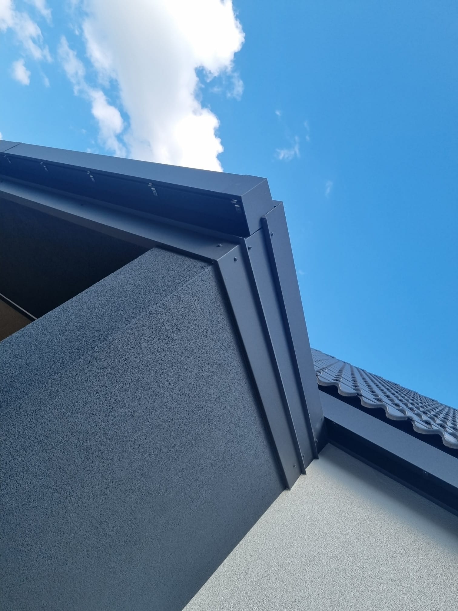 Szczegółowe ujęcie narożnika domu z szarą elewacją, ciemnoszarym obramowaniem i fragmentem dachu pokrytego szarą dachówką na tle błękitnego nieba z białymi obłokami.