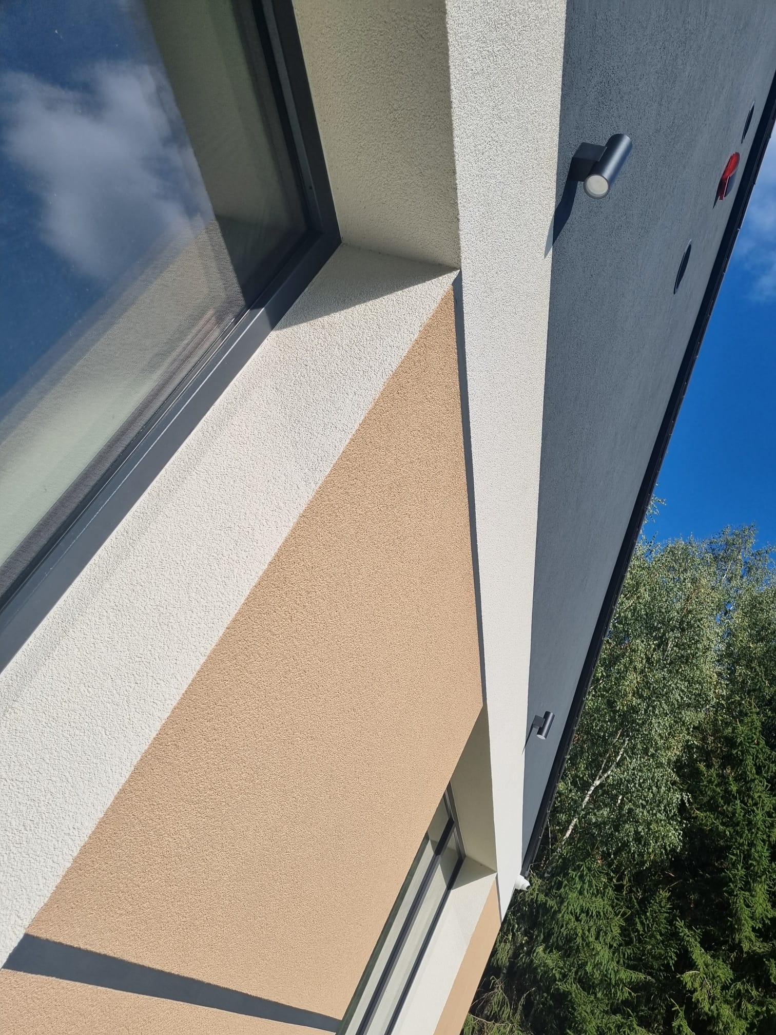 Fragment elewacji budynku z widocznym oknem, oświetleniem zewnętrznym i fragmentem zieleni w tle; elewacja w odcieniach beżu, bieli i szarości.