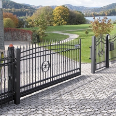 Otwarta, czarna, metalowa brama wjazdowa z dekoracyjnymi elementami, prowadząca na posesję wyłożoną kostką brukową, w tle zielony trawnik, drzewa i jezioro.