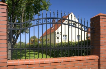 Czarne, ozdobne ogrodzenie metalowe z ceglanymi słupkami, widoczne na tle domu z czerwoną dachówką i zielenią drzew.