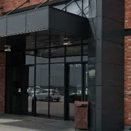 Wejście do perfumerii z oznakowaniem dla osób niepełnosprawnych, fasada z cegły i czarnych paneli, widoczne kosze na śmieci.