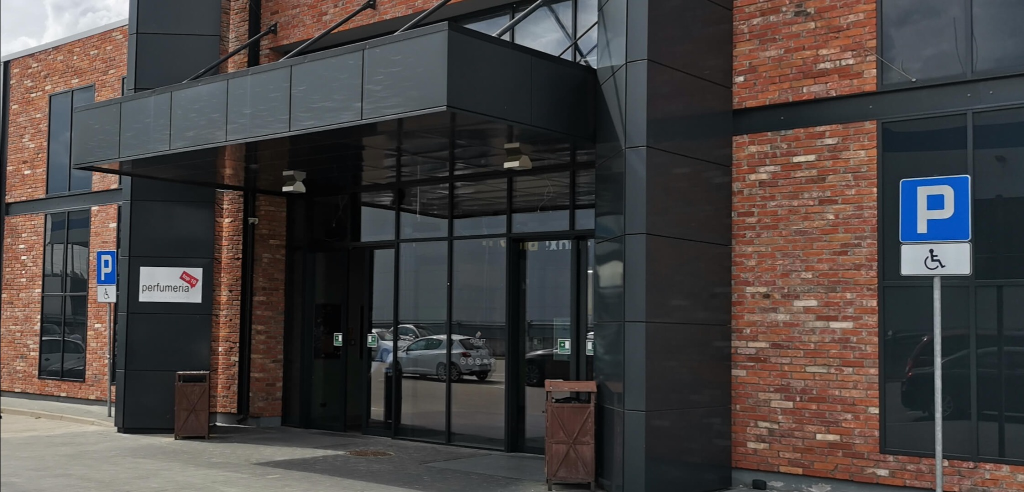Wejście do perfumerii z oznakowaniem dla osób niepełnosprawnych, fasada z cegły i czarnych paneli, widoczne kosze na śmieci.