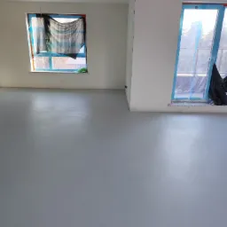 Szara, gładka posadzka w jasnym pokoju z zabezpieczonymi oknami. Widoczne detale wykończenia i przygotowania do dalszych prac wykończeniowych.