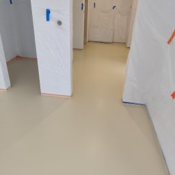 Mix bud Maciej Pawłowski - Jasna, beżowa posadzka w pomieszczeniu zabezpieczonym folią malarską. Widoczne kolumny i ściany przygotowane do dalszych prac wykończeniowych. Minimalistyczny kadr.