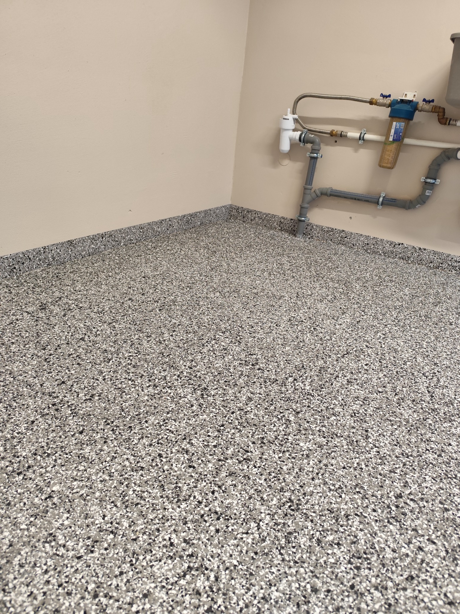 Szara podłoga z żywicy epoksydowej z widocznymi rurami i filtrami w rogu pomieszczenia.