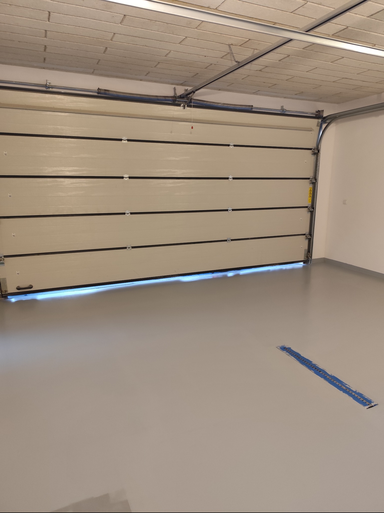 Gładka, szara podłoga w garażu z segmentową bramą garażową w kolorze ecru, z widoczną listwą uszczelniającą u dołu, niebieska taśma malarska na podłodze, sufit z betonu komórkowego.