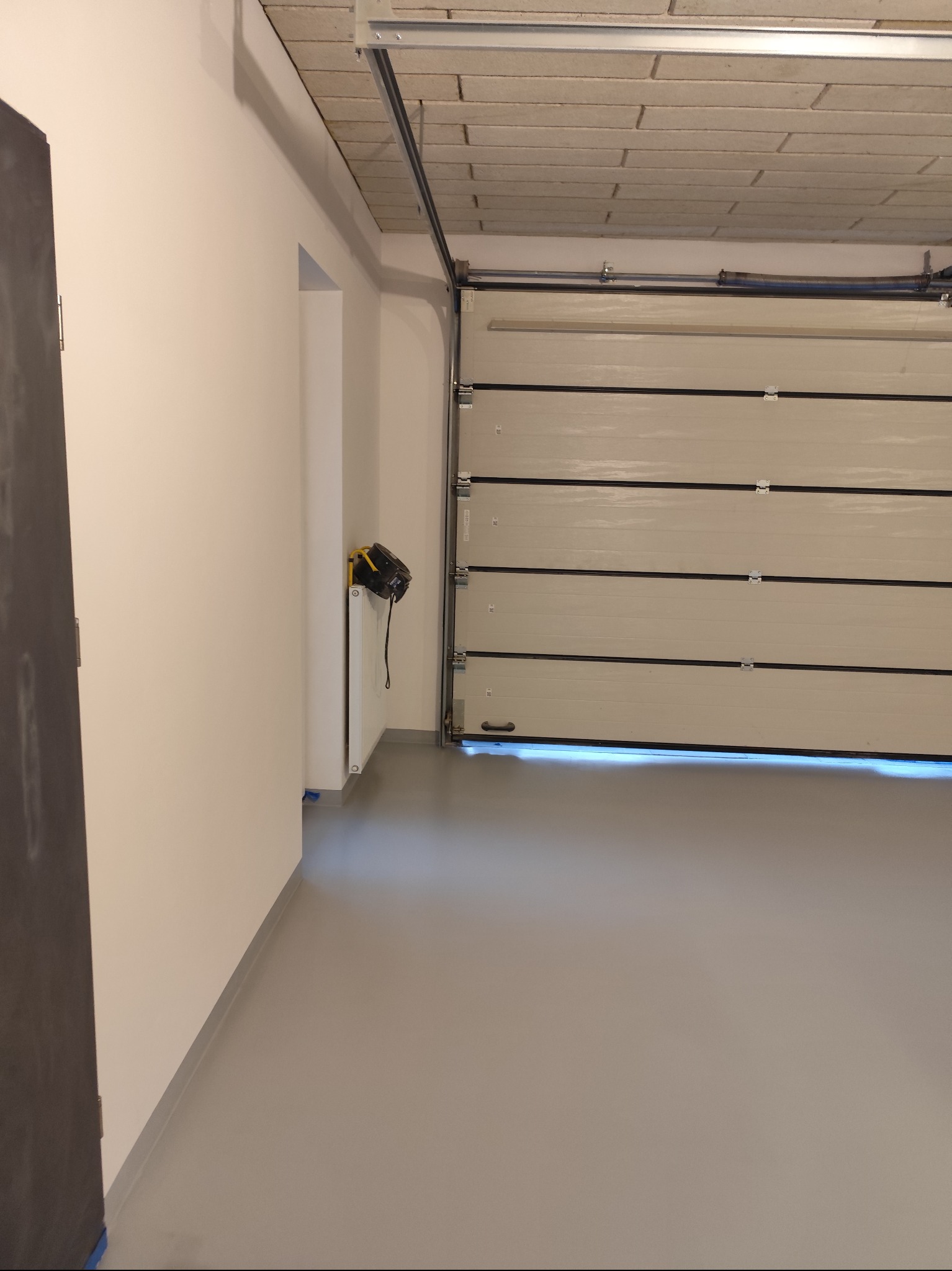 Szara, gładka podłoga w garażu z segmentową bramą garażową i lampą budowlaną na grzejniku w tle.