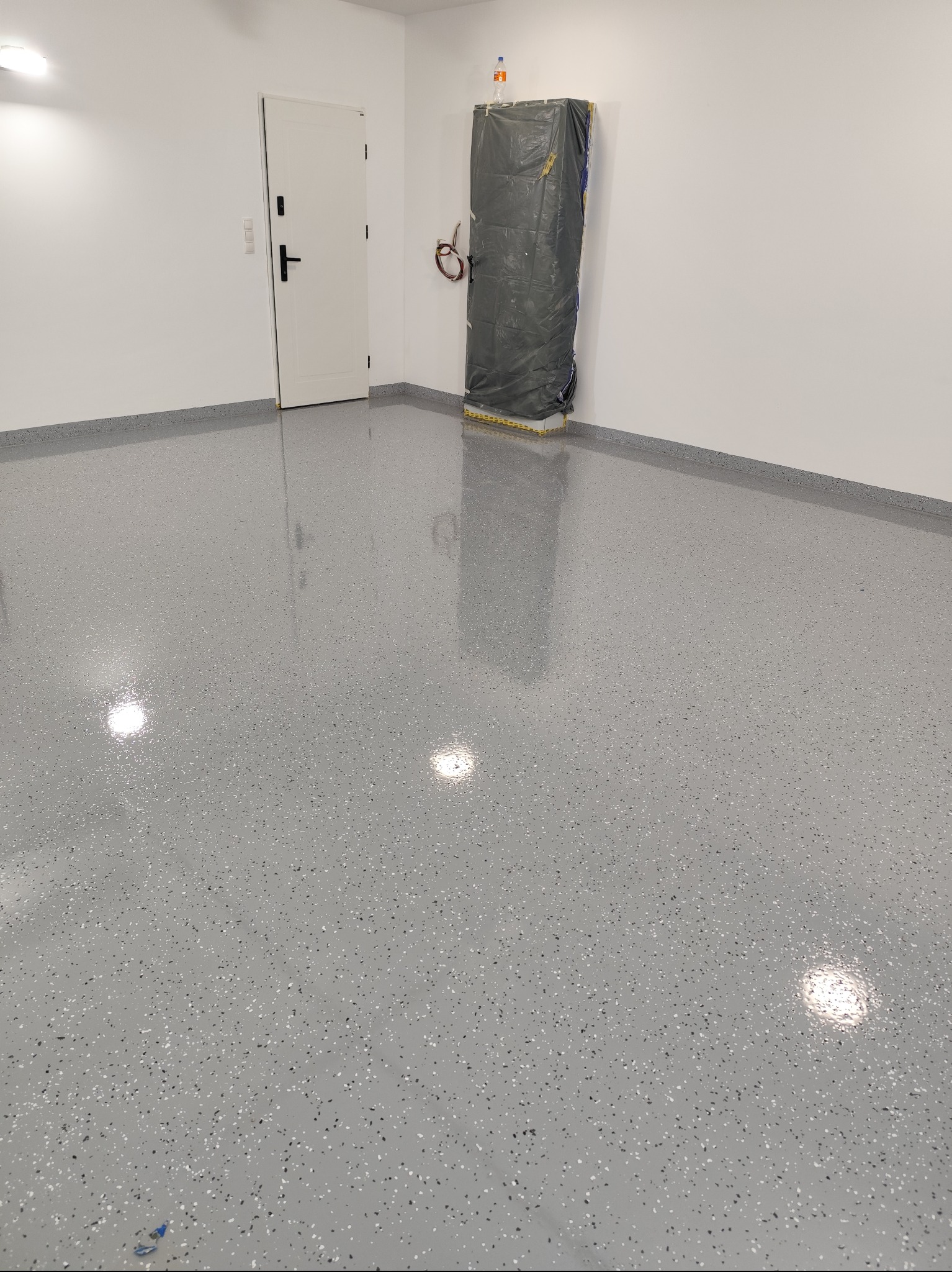 Szara, błyszcząca posadzka żywiczna z drobnymi, kolorowymi wtrąceniami w nowym garażu, zabezpieczona folią ściana w tle, białe drzwi po lewej stronie.
