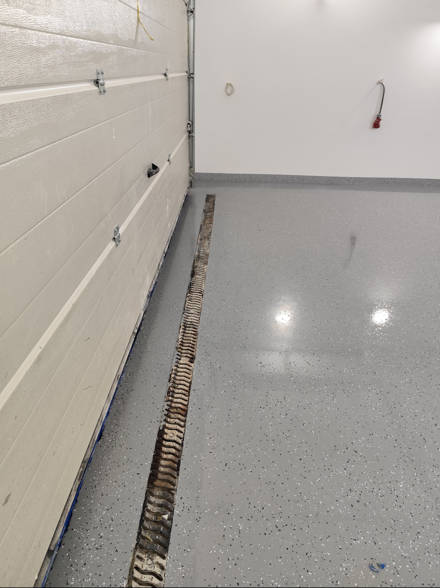 Szara, błyszcząca posadzka z drobnymi, kolorowymi wtrąceniami w garażu w Warszawie, z widocznym odpływem liniowym i fragmentem bramy garażowej.