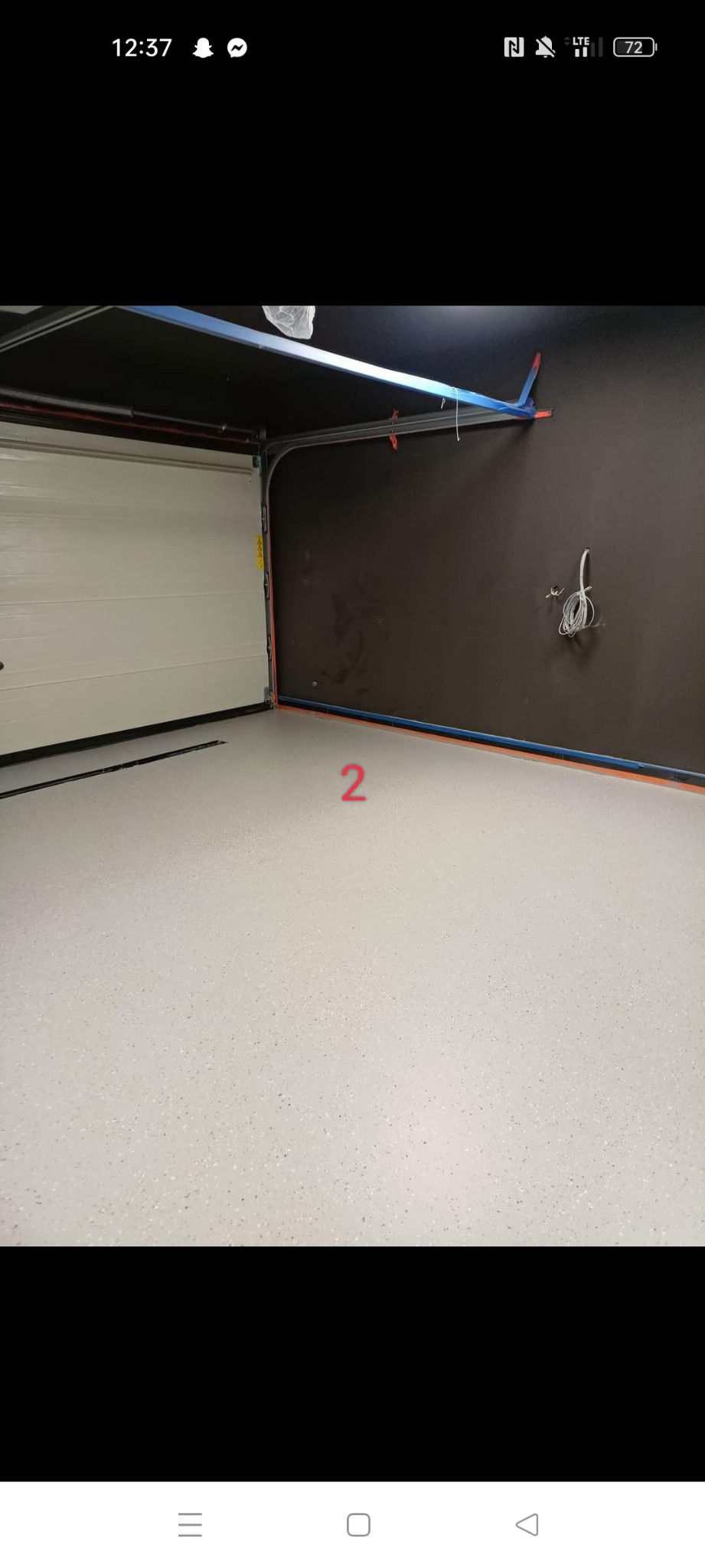 Jasnoszara posadzka w garażu z zamkniętą białą bramą segmentową i ciemnoszarą ścianą, widoczne przewody elektryczne.