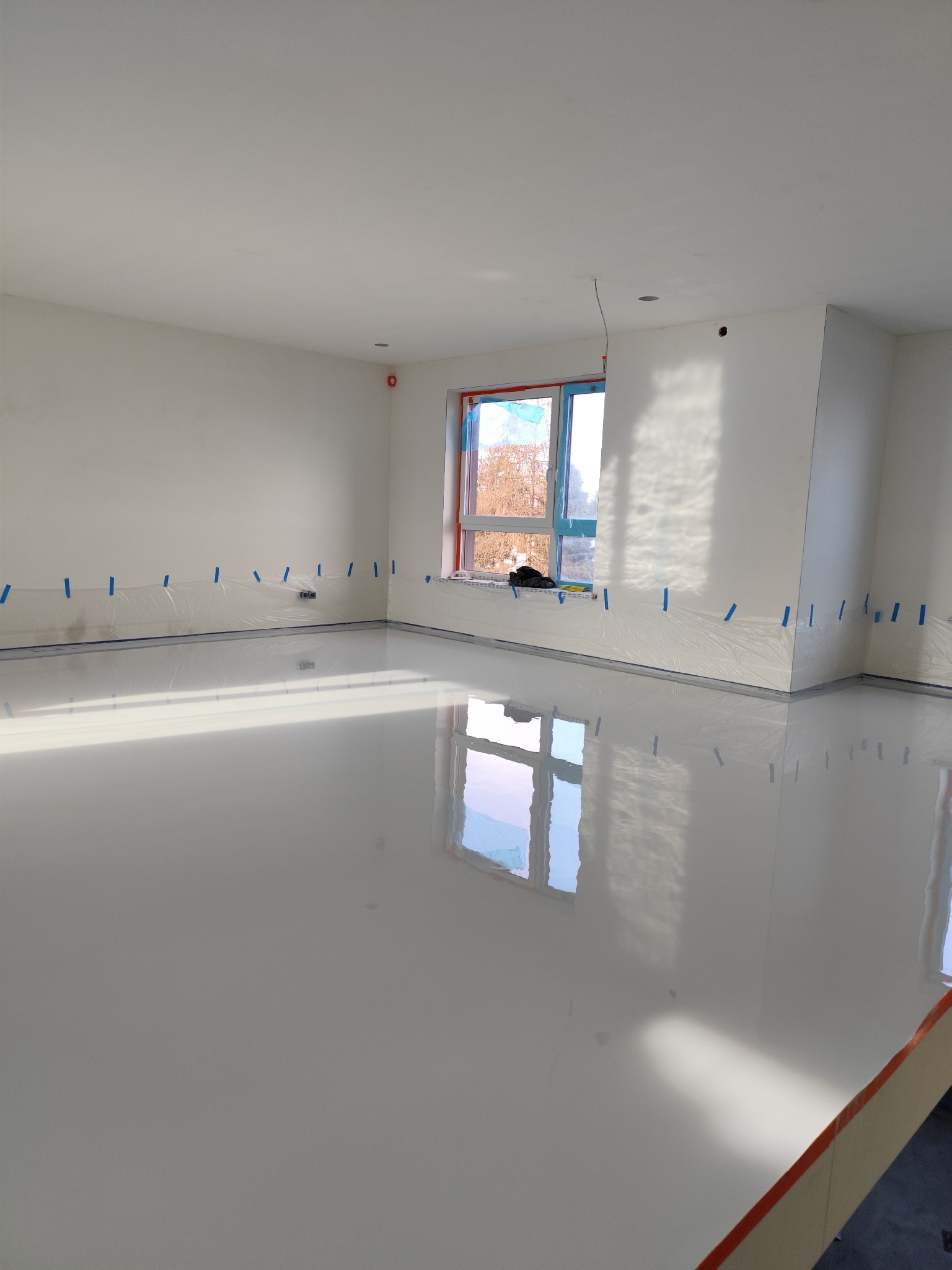 Świeżo wylana, jasna posadzka w pomieszczeniu z oknem, zabezpieczone ściany folią i niebieską taśmą malarską.