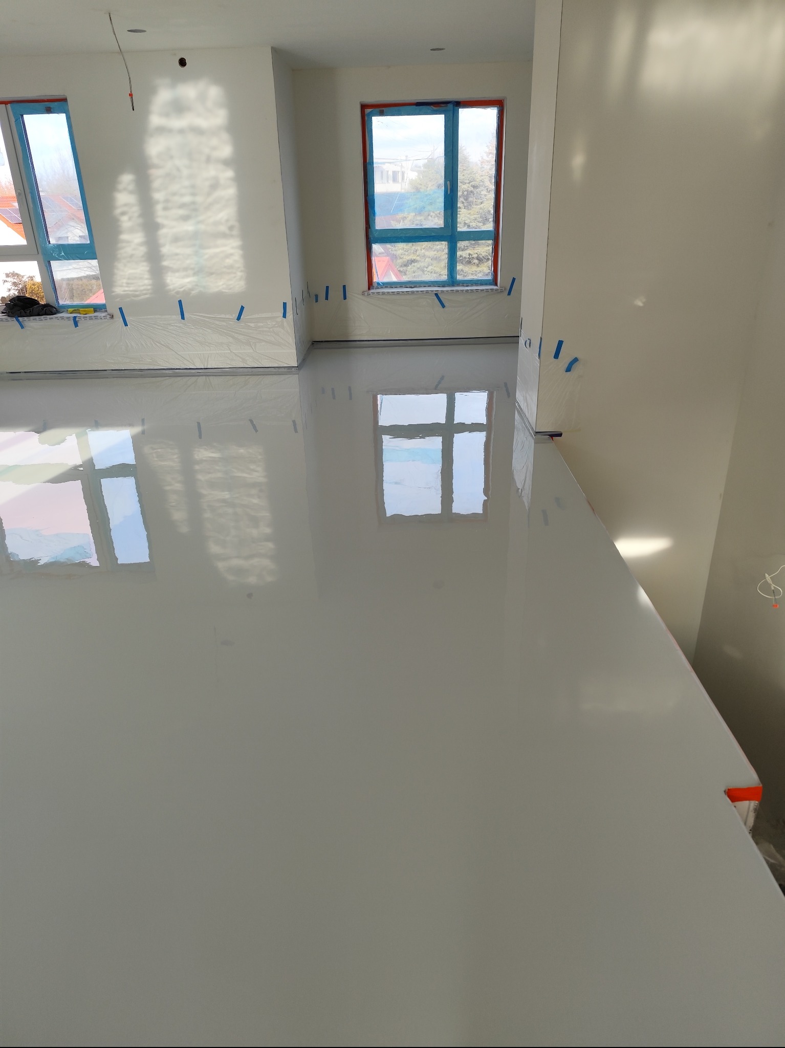 Jasna, gładka podłoga w pomieszczeniu z oknami, zabezpieczonymi folią i taśmą malarską, odbijająca światło i widok z okien.