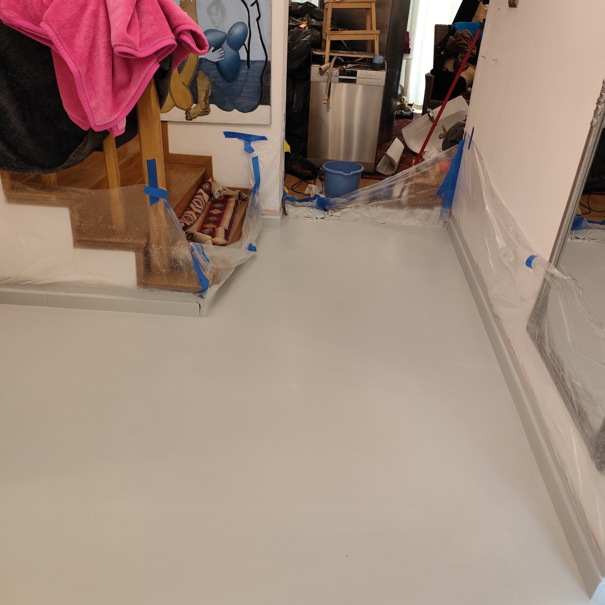 Świeżo wylana, jasnoszara podłoga w pomieszczeniu mieszkalnym, zabezpieczona folią i niebieską taśmą malarską przy schodach i ścianach, z widocznym fragmentem obrazu na ścianie i sprzętami AGD w tle.