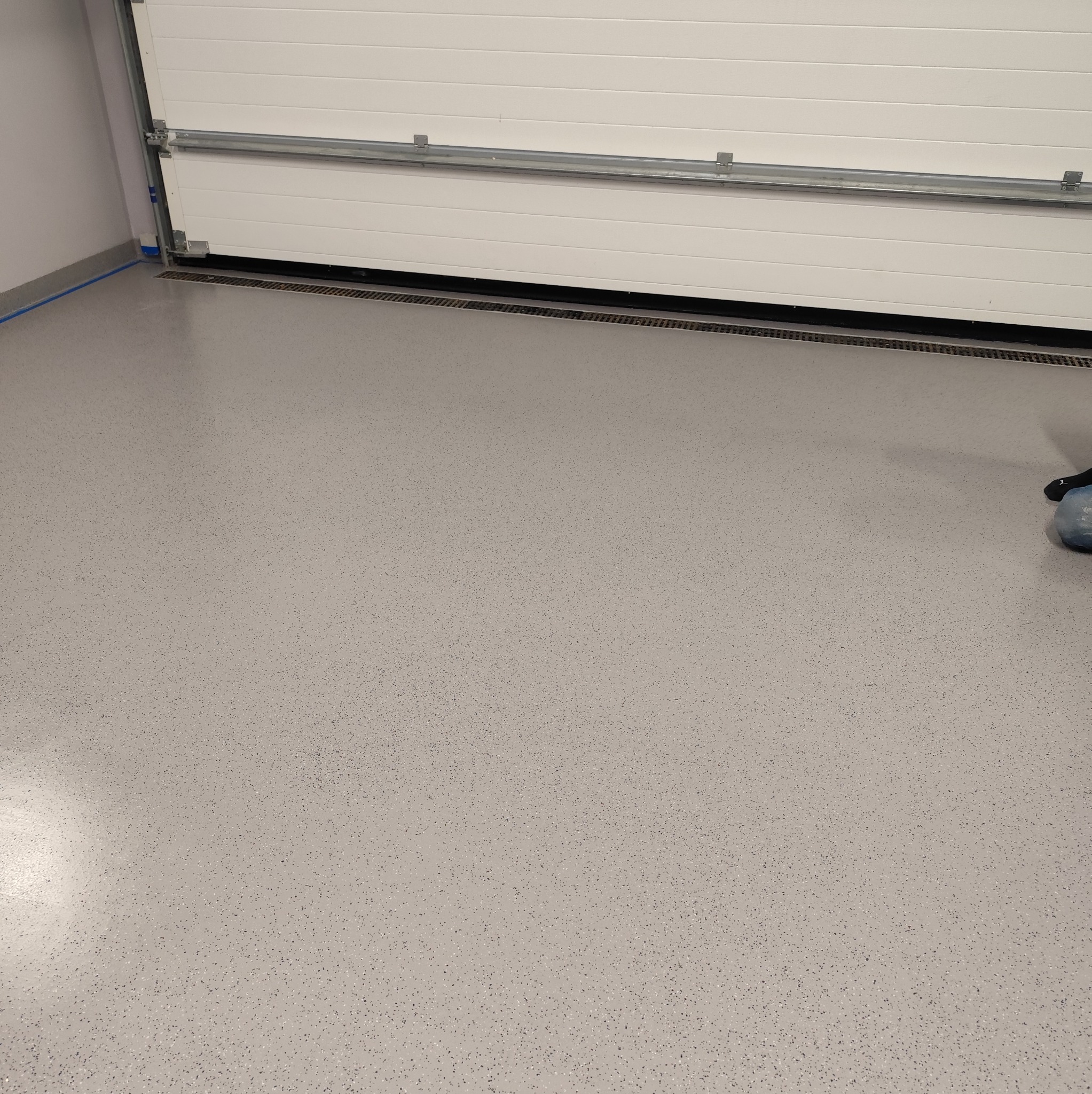 Jasnoszara, gładka posadzka z delikatnym wzorem ziarnistym, widoczny fragment bramy garażowej i odpływ liniowy przy ścianie.