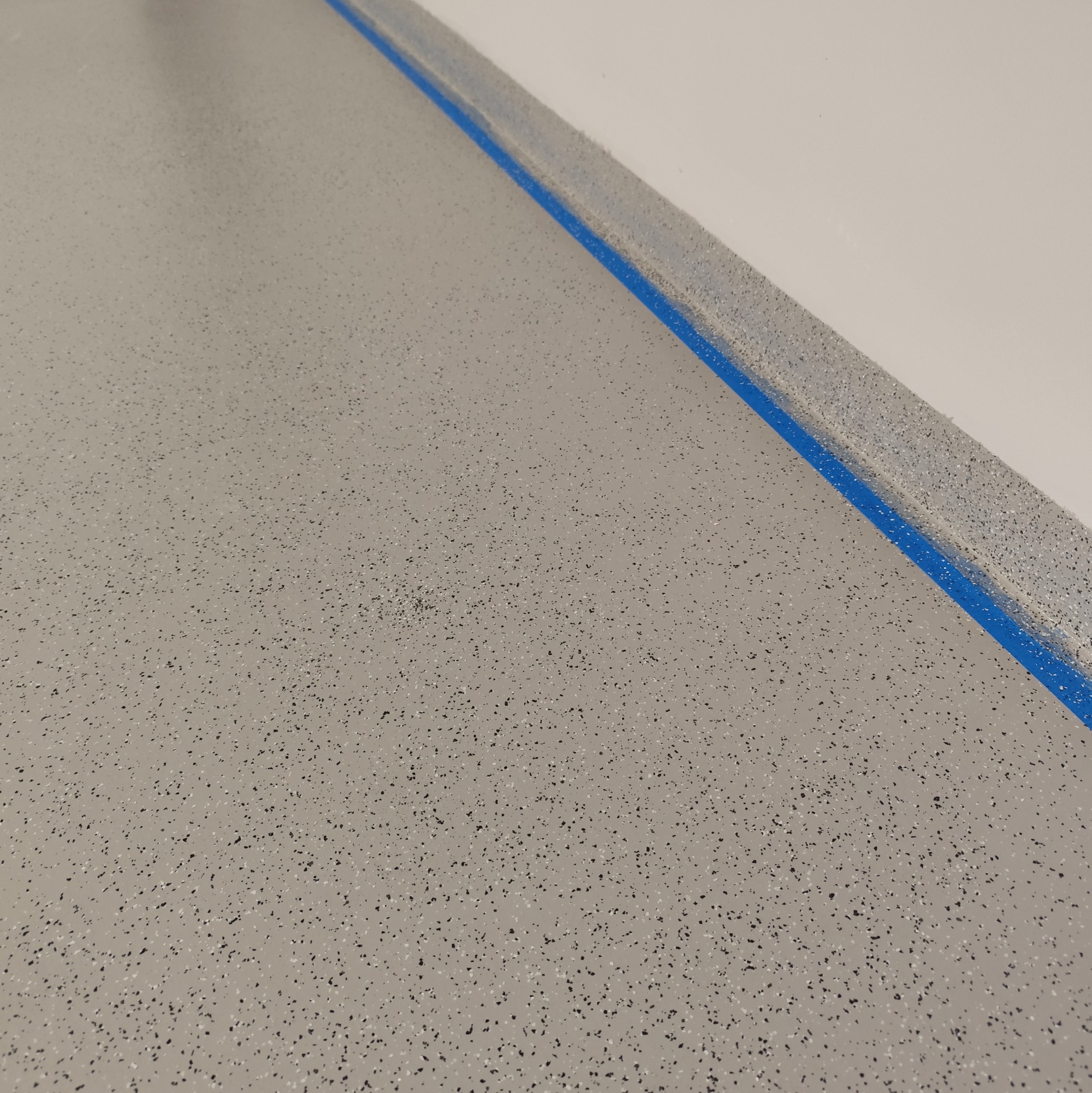 Szara posadzka z drobnymi czarnymi i białymi elementami, zabezpieczona niebieską taśmą malarską przy ścianie.