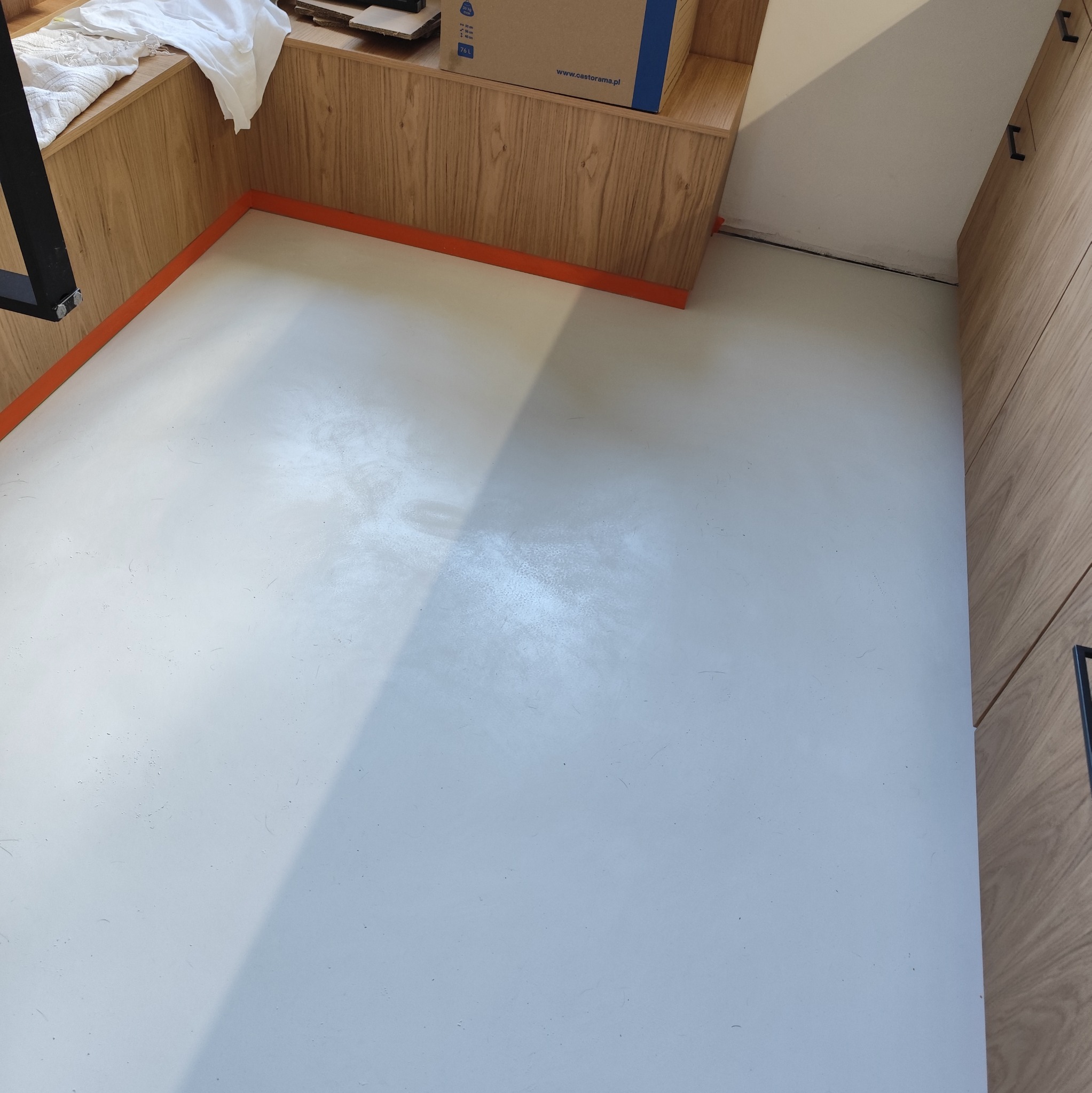 Jasnoszara, gładka posadzka żywiczna w pomieszczeniu z drewnianą zabudową, zabezpieczona pomarańczową listwą przyścienną.