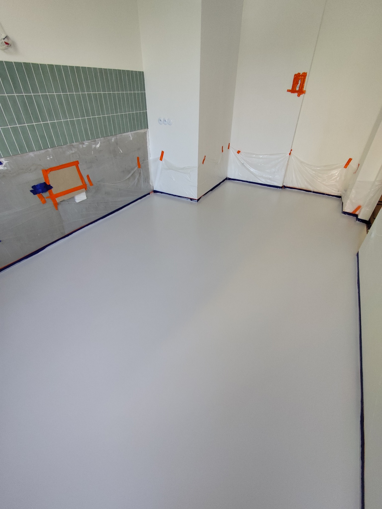 Jasnoszara, gładka podłoga w trakcie prac wykończeniowych, zabezpieczona folią i taśmą malarską przy ścianach z szarymi płytkami.