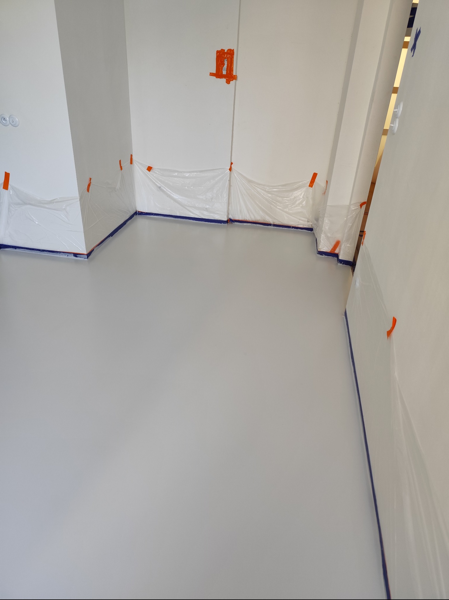 Świeżo wylana, jasnoszara podłoga w pomieszczeniu, zabezpieczona folią malarską przy ścianach, widoczne pomarańczowe taśmy mocujące folię.