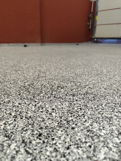 Szara posadzka z żywicy epoksydowej z czarnymi i białymi elementami, wykończona w garażu z częściowo otwartą bramą.