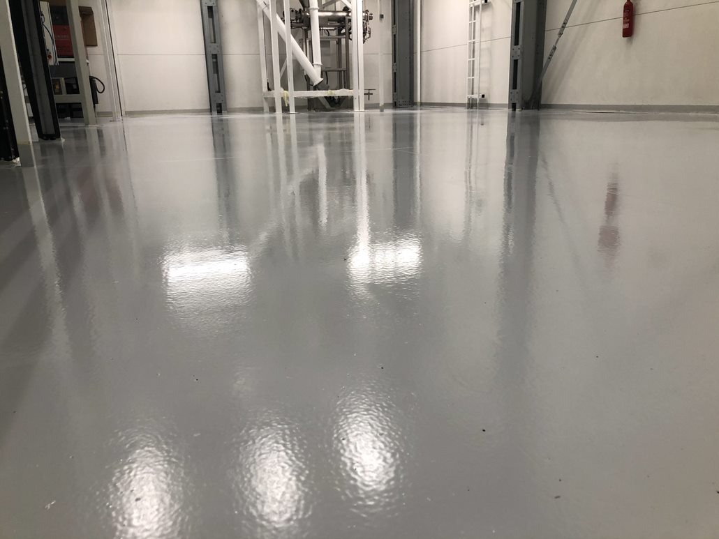 Błyszcząca, szara powierzchnia przemysłowa. Gładka, żywiczna podłoga z refleksami świetlnymi w hali produkcyjnej z elementami konstrukcyjnymi w tle.