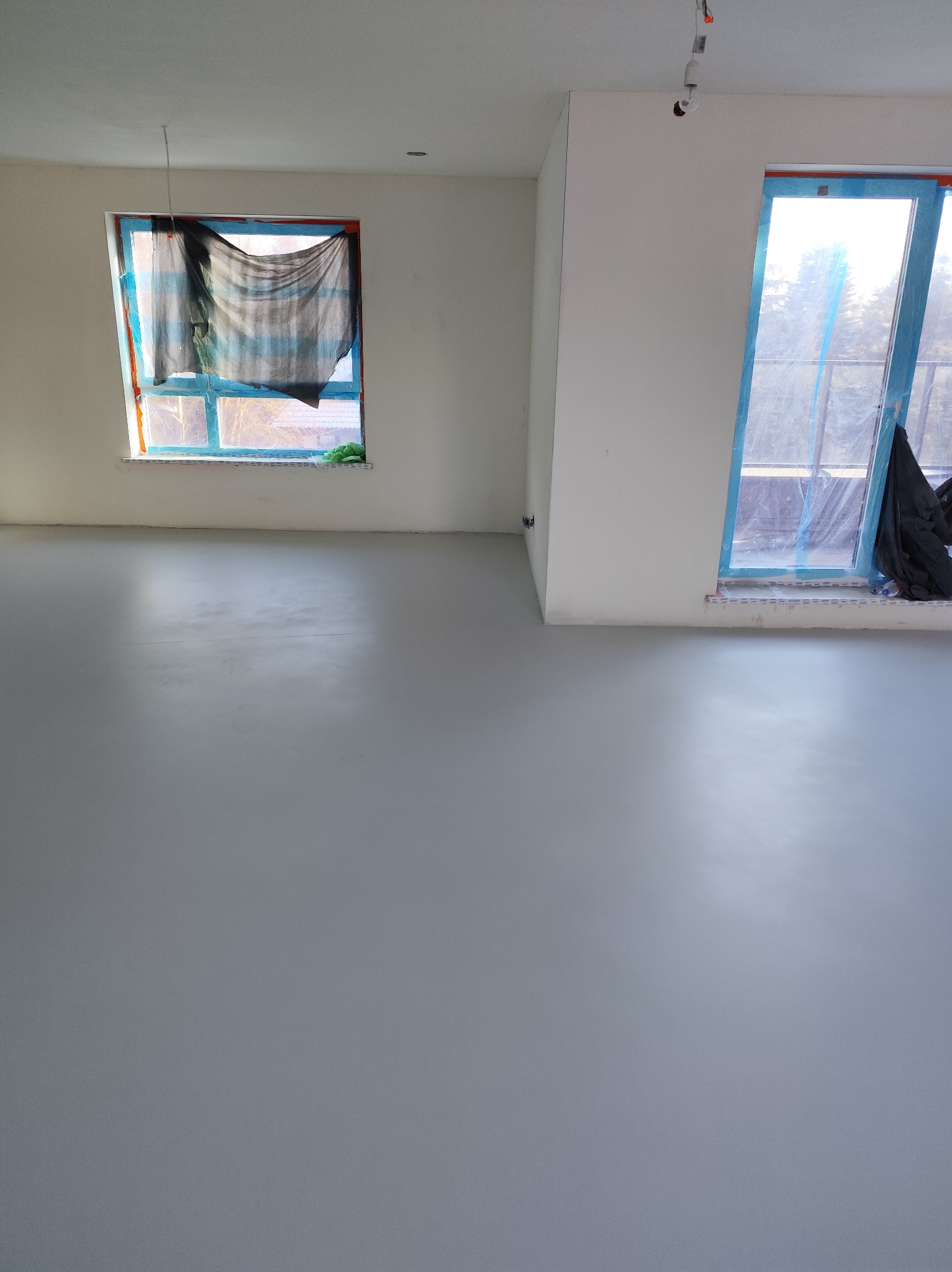 Szara, gładka posadzka w jasnym pokoju z zabezpieczonymi oknami. Widoczne detale wykończenia i przygotowania do dalszych prac wykończeniowych.