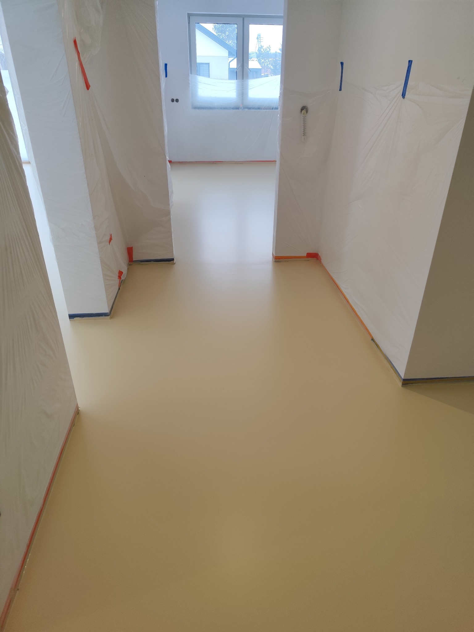 Beżowa, gładka podłoga w jasnym wnętrzu, zabezpieczone folią ściany i filary. Widok na okno w głębi. Minimalistyczny design, świeżo wylana powierzchnia.