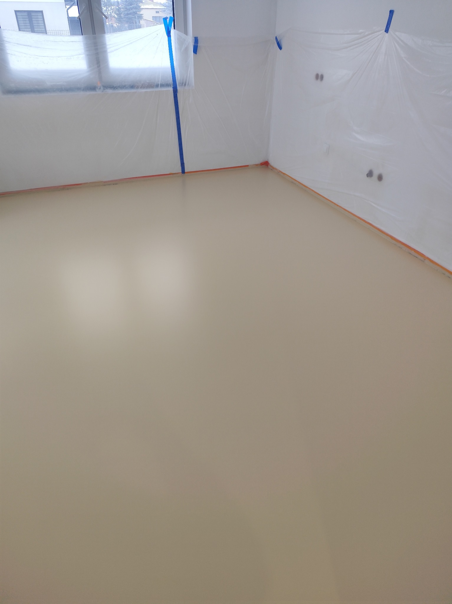 Beżowa, gładka powierzchnia wylanej podłogi w pokoju zabezpieczonym folią malarską. Widoczne taśmy malarskie i gniazdka elektryczne.