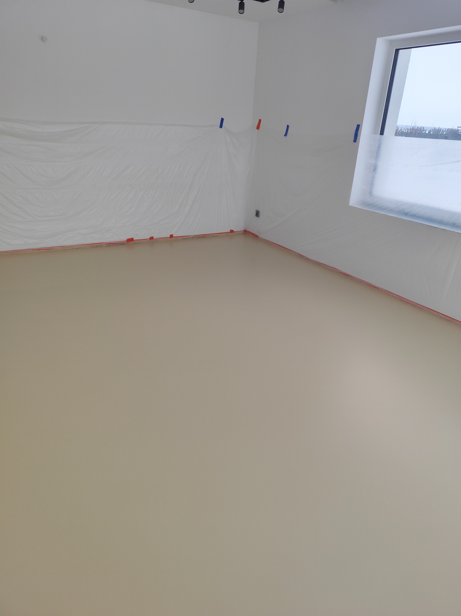 Jasna, gładka podłoga w nowym wnętrzu, zabezpieczona folią malarską przy ścianach i oknach. Minimalistyczny design, przygotowanie do dalszych prac wykończeniowych.